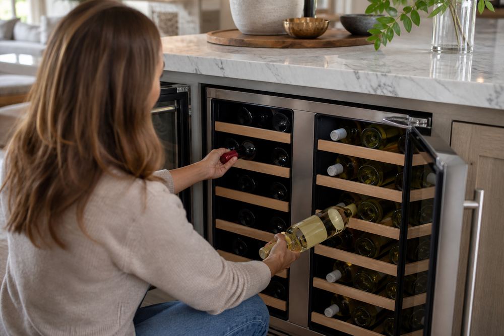 Open dual-zone wine cooler under a quartz kitchen island, reds and whites on separate shelves, host’s hand selecting a dinner bottle.