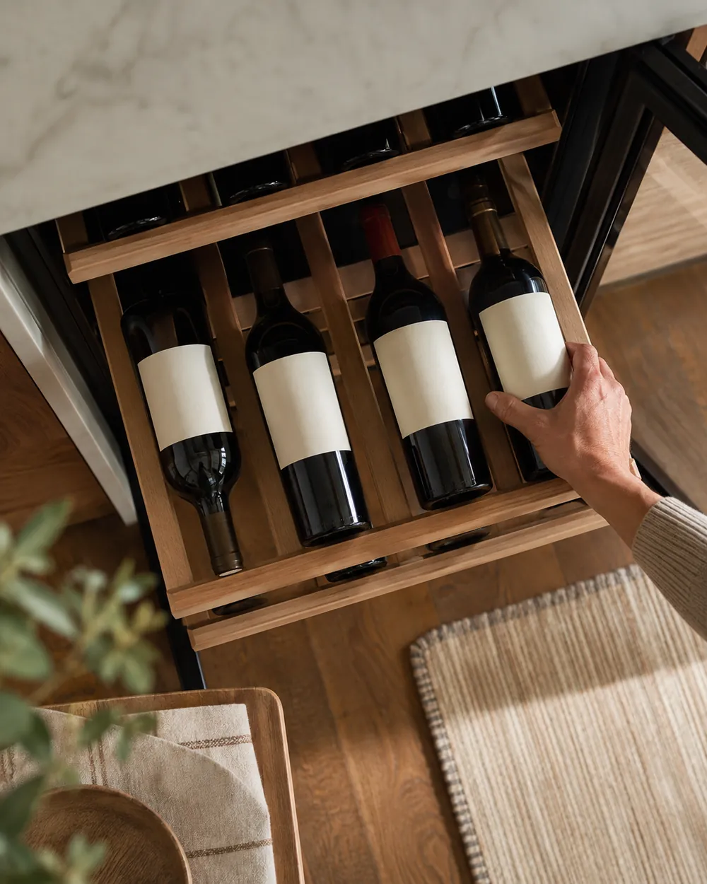 One hand adjusts staggered wine bottles on wooden cooler shelves, spacing labels upward to prevent scraping and rattling, overhead view.