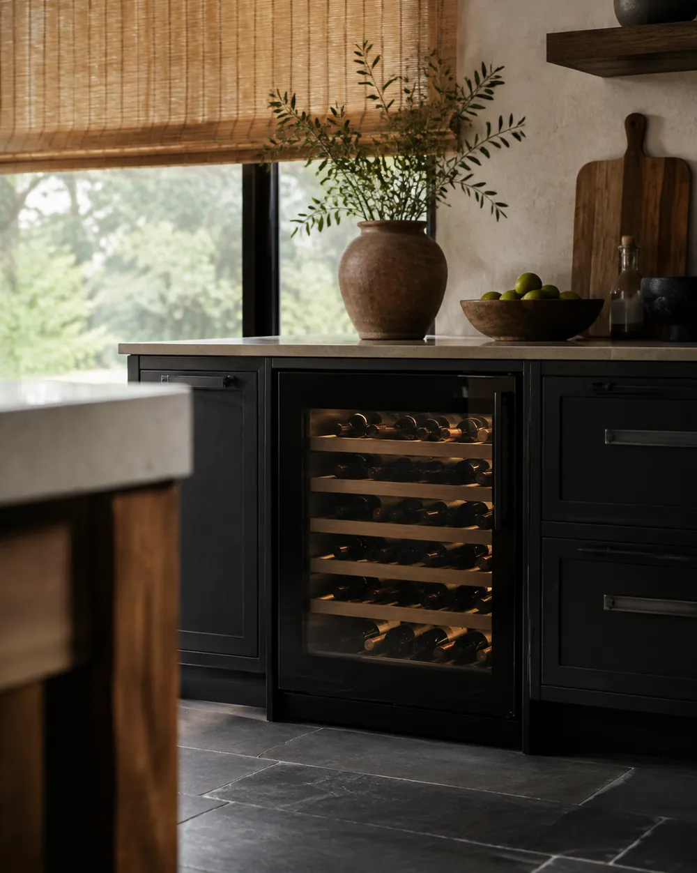 Stone tile flooring foregrounds a low-angle view of a wine cooler tucked beside shaded cabinets, away from sunlit windows.